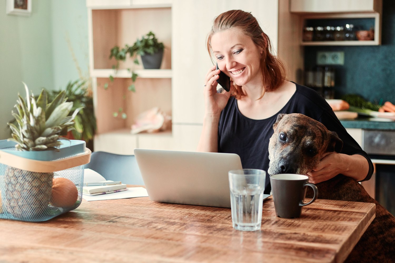 A smiling woman on the phone at her kitchen table, laptop open, her dog resting its head beside her