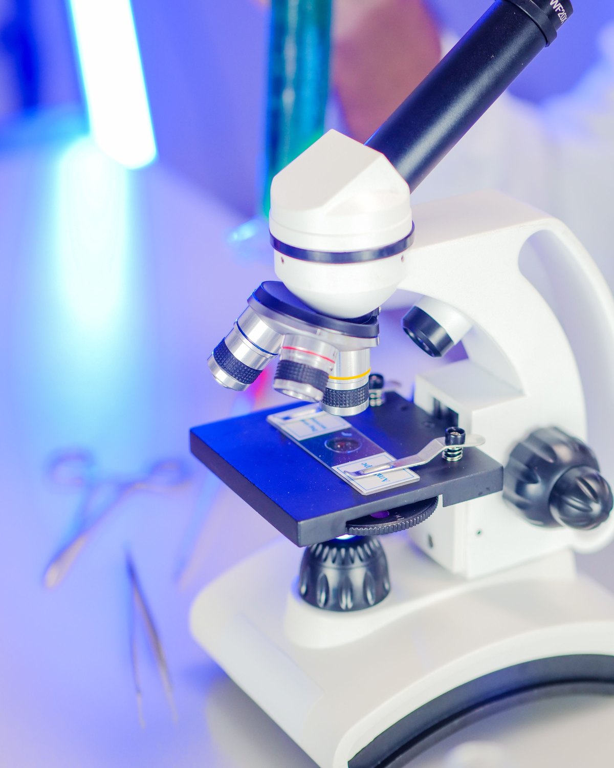 A white laboratory microscope with a glass slide on its stage, lit in cool blue light