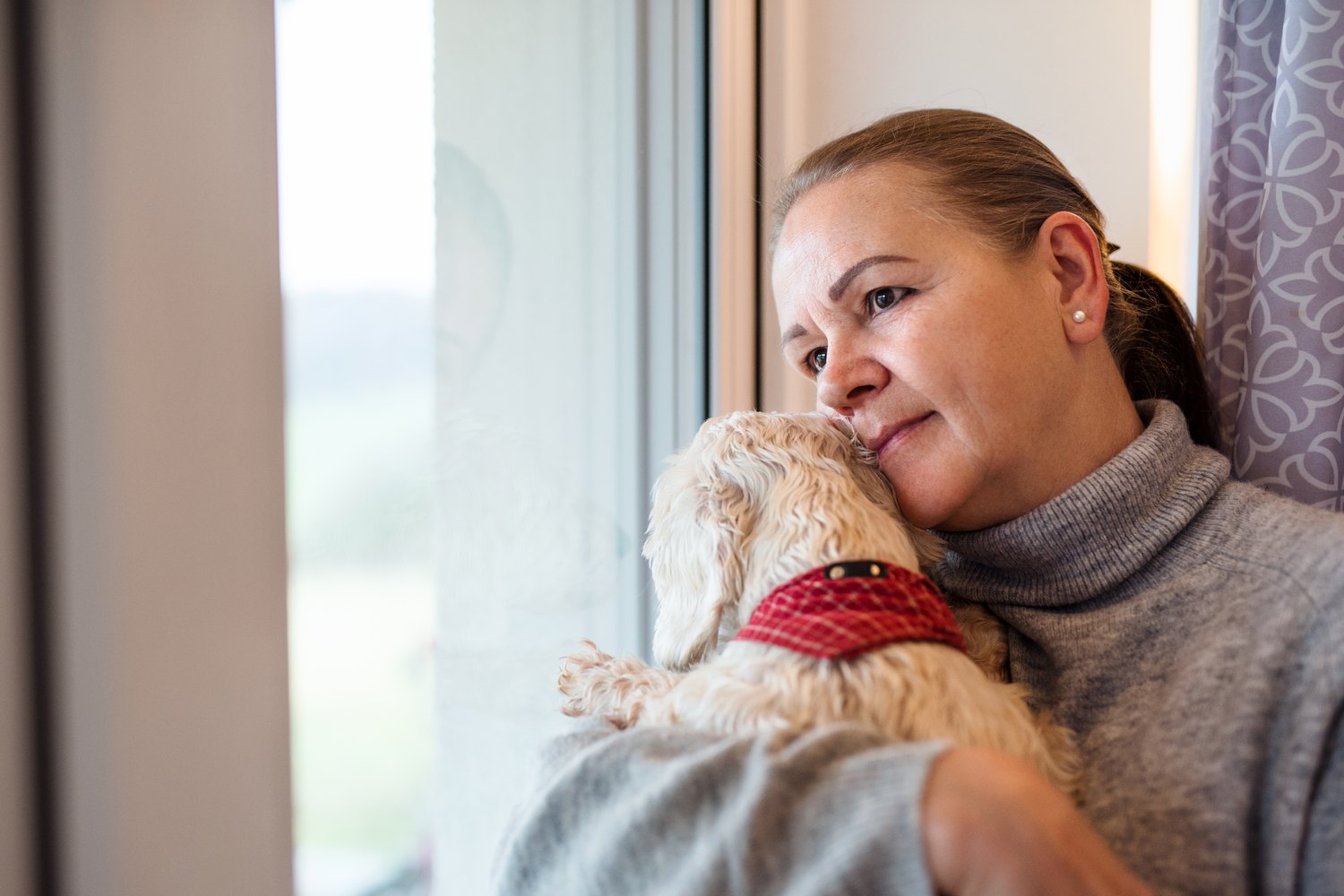 A woman by a window holding her small white dog close, cheek to cheek