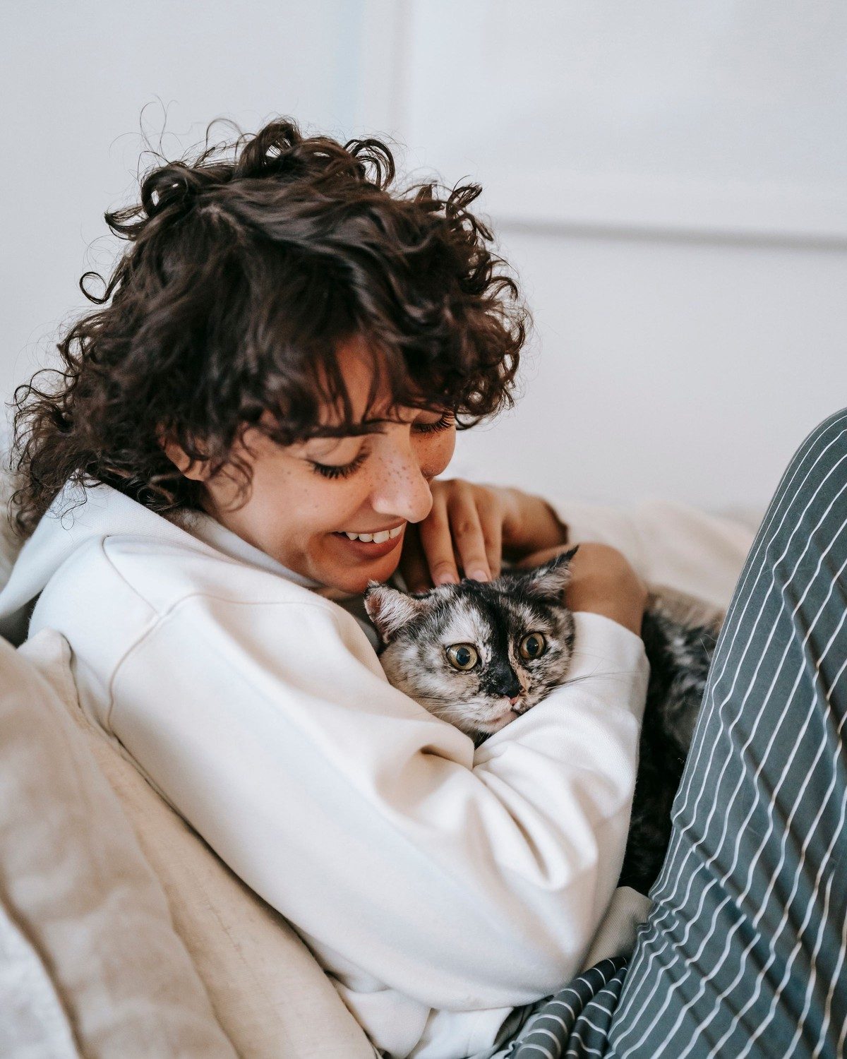 A smiling woman curled up on a sofa, hugging her tortoiseshell cat close