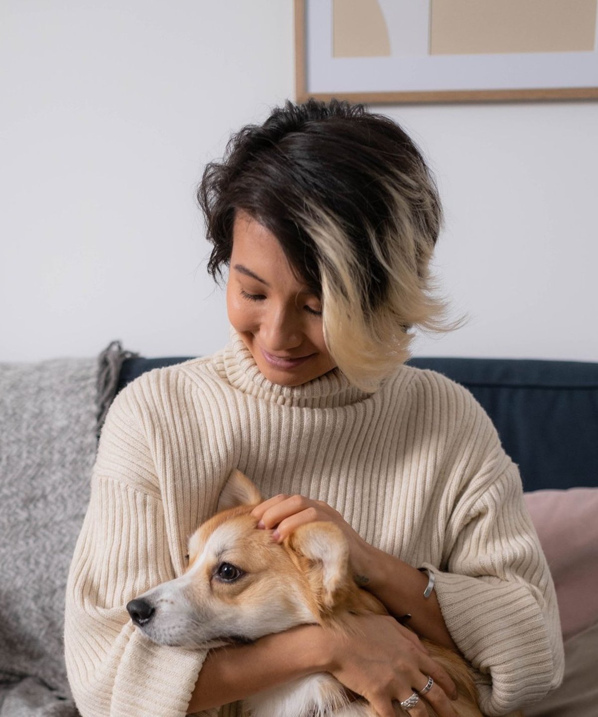A woman on a sofa smiling down at the corgi she holds, her hand resting on its head